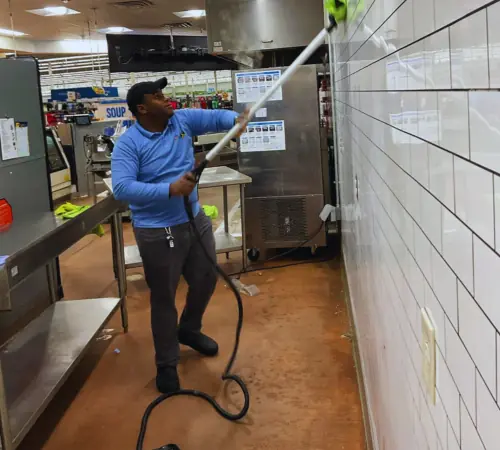Commercial kitchen tile cleaning in a grocery backroom, technician scrubbing white subway tiles with long-handled scrubber