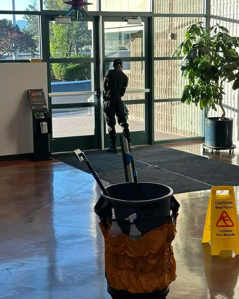 Janitorial cleaner opening a glass commercial lobby door with mop bucket, caution sign, and entrance mat visible