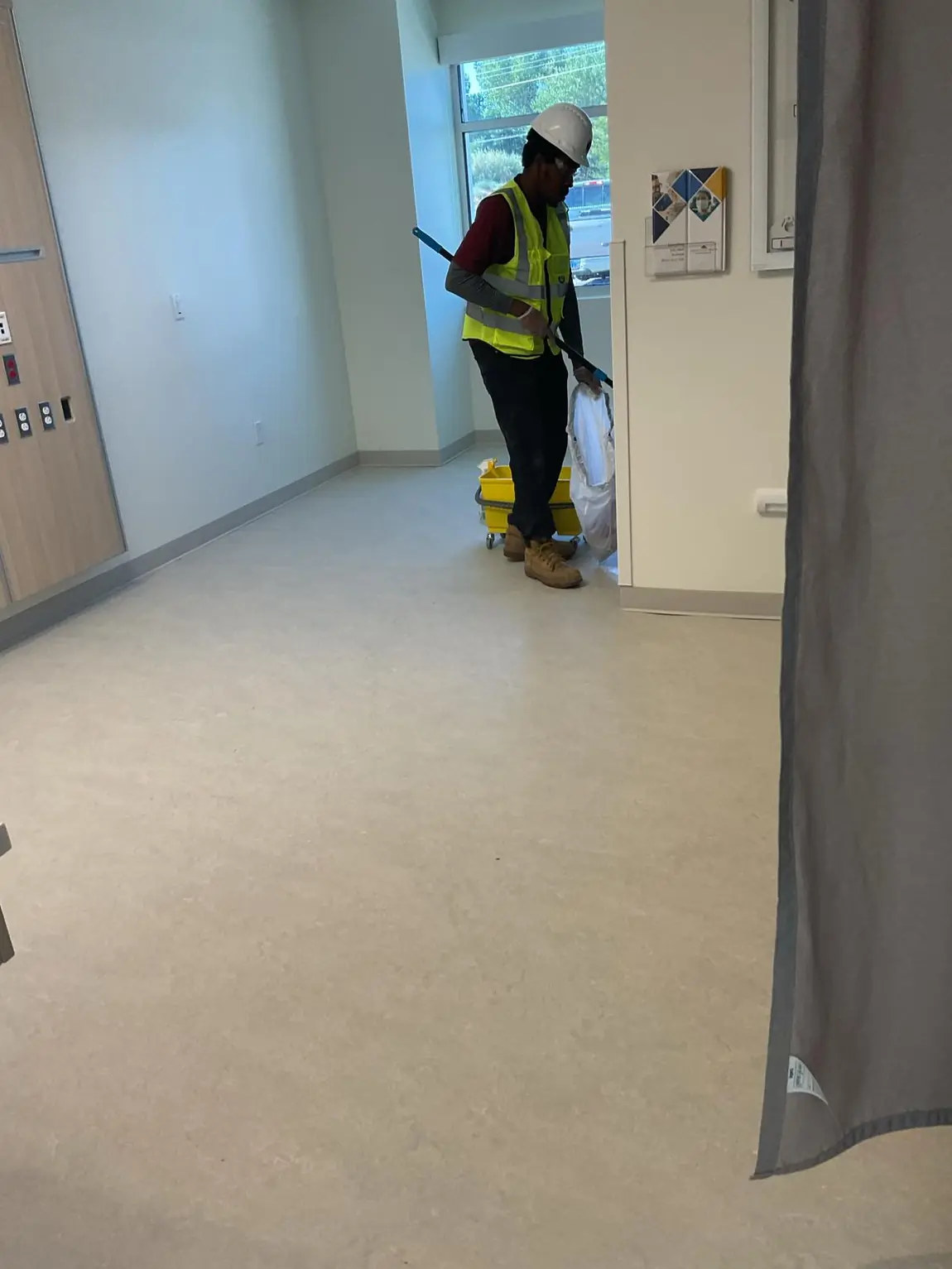 Janitorial worker in hard hat and vest mopping a clean medical facility room with cart and supplies