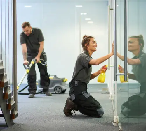 Two professional commercial cleaners cleaning an office: one kneeling wiping glass door, another vacuuming carpet