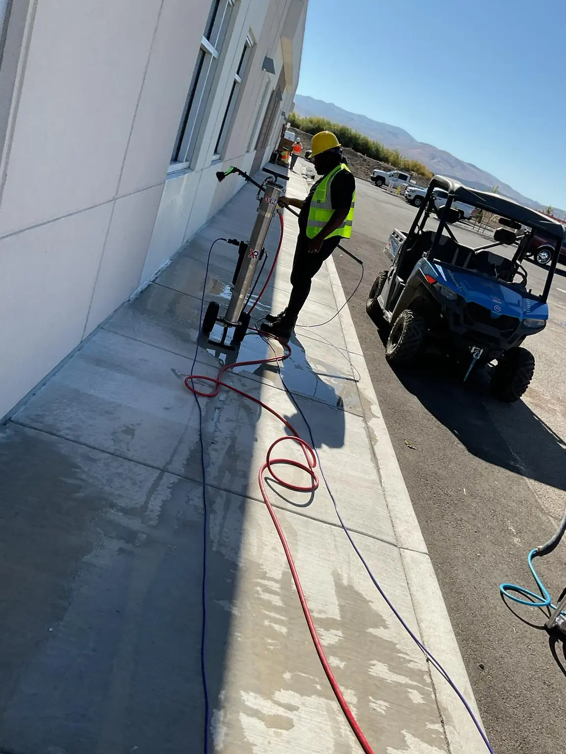 Worker in hard hat and hi-vis vest pressure-washing exterior concrete sidewalk during commercial post-construction cleaning