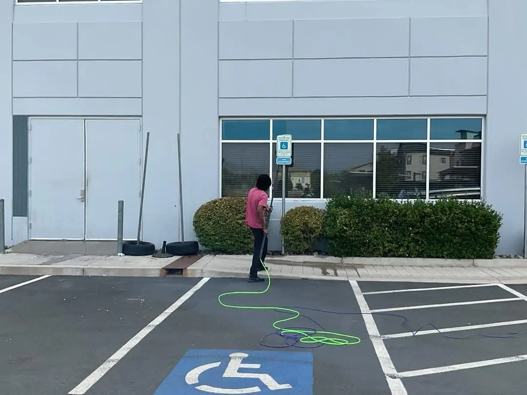 Worker pressure washing a commercial building exterior near storefront windows and handicap parking in a business plaza