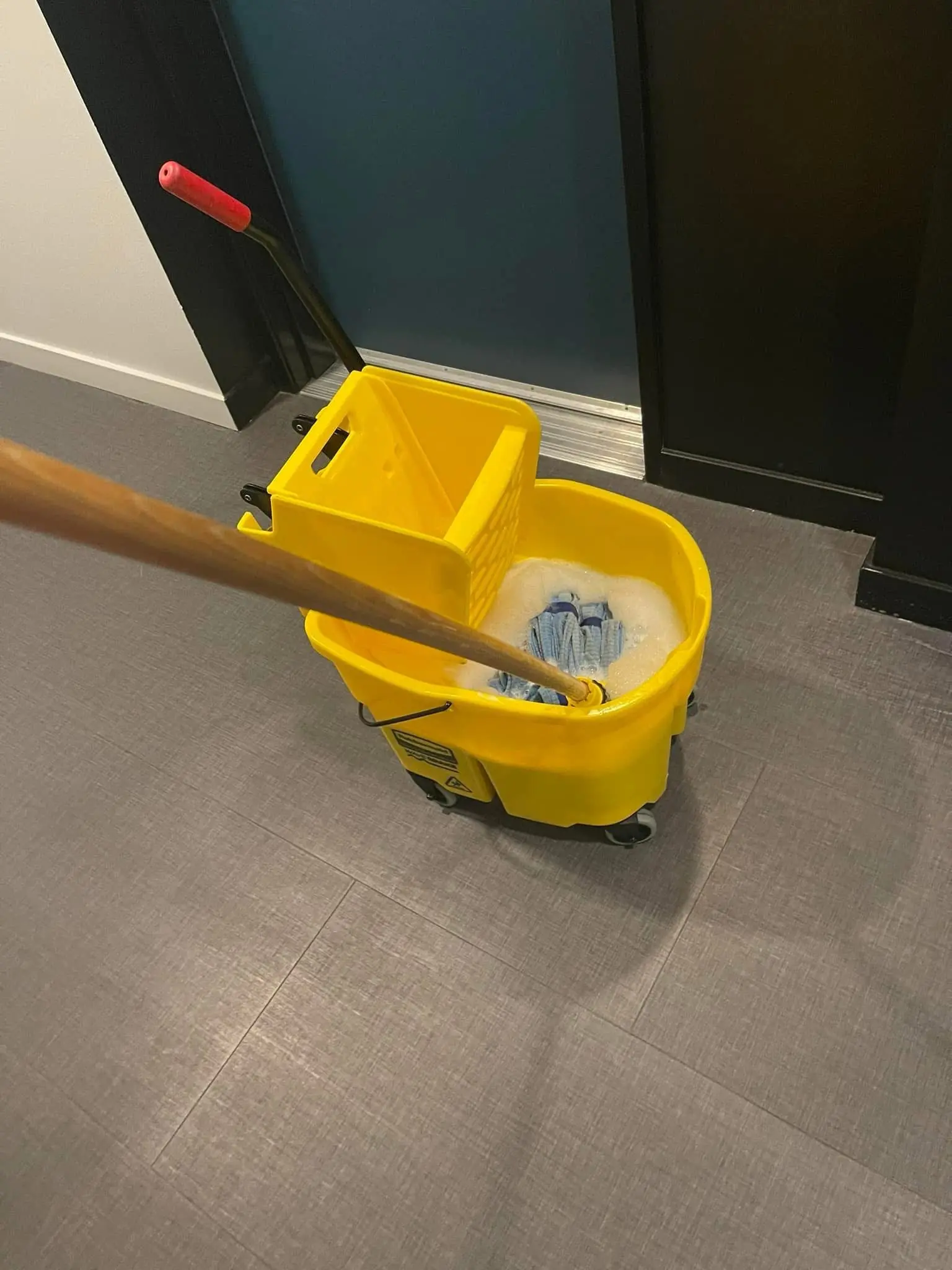 Yellow mop bucket with wringer and soapy mop in a commercial hallway on gray tile flooring for janitorial cleaning